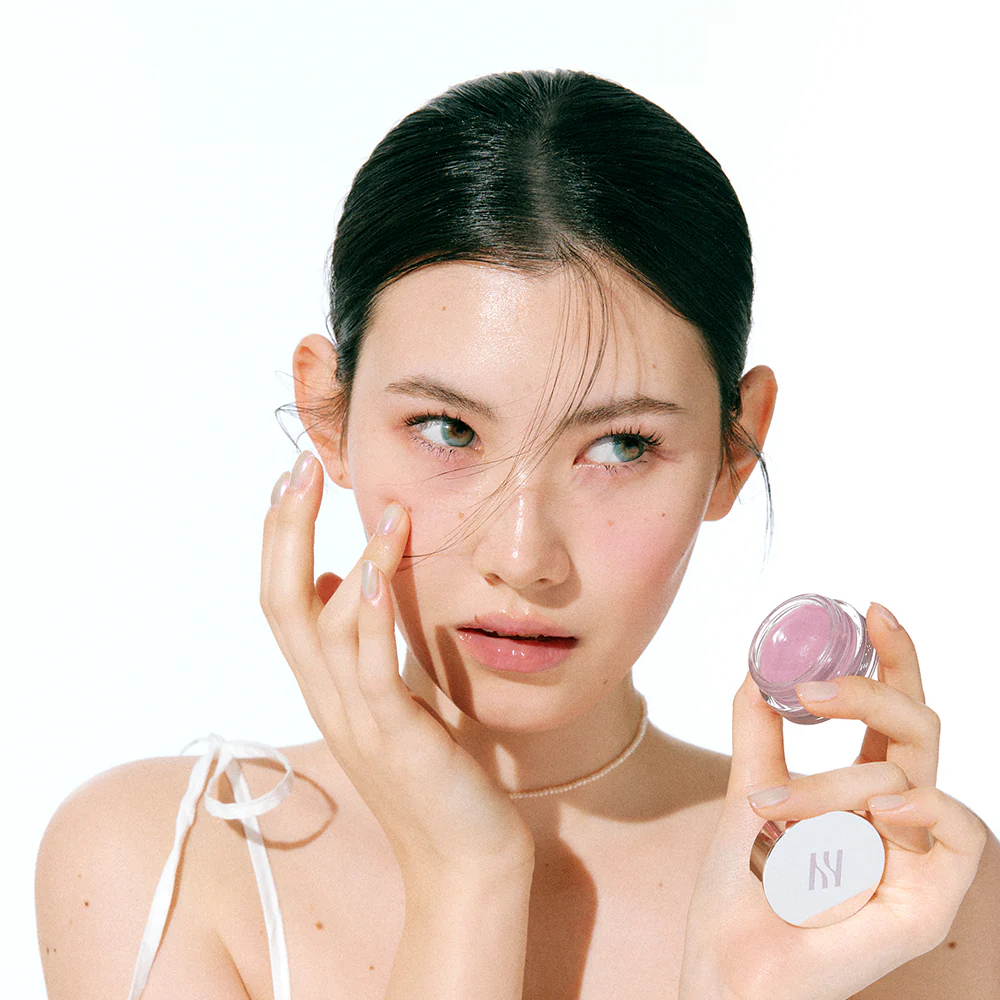 HERA Sensual Lip & Cheek 7g : maquillage corail sur visage lumineux, modèle appliquant avec le produit HERA, fond clair.