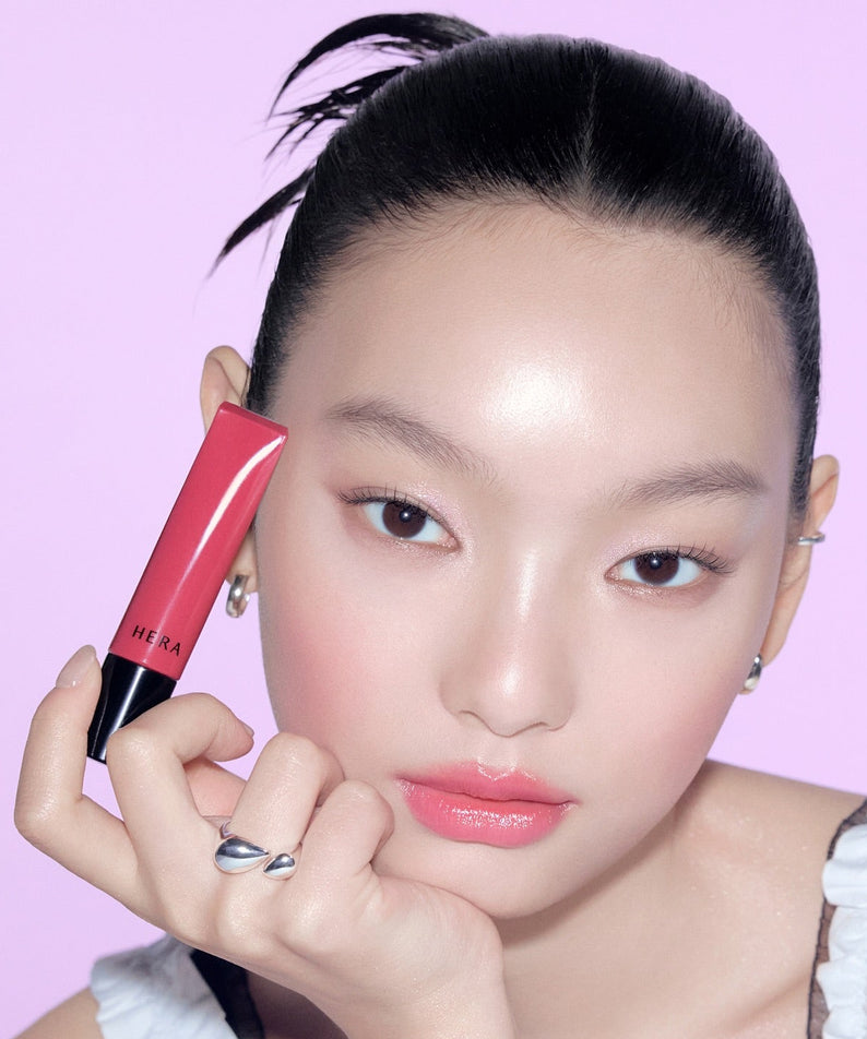 HERA Sensual Tinted Shine Lip Serum 8.5g de HERA, flacon rose tenu par un mannequin sur fond violet.