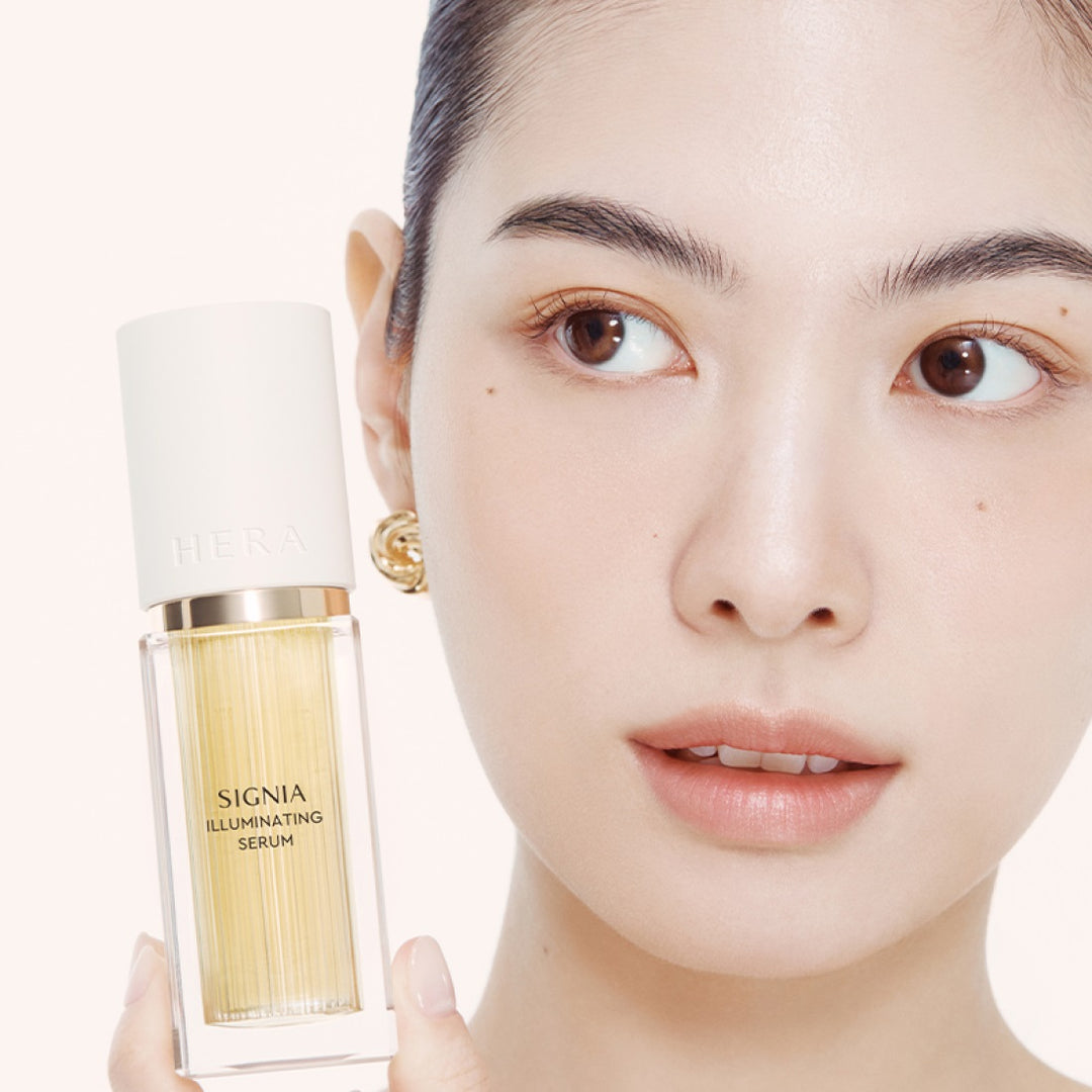 Décris l'image en français : HERA  Signia Illuminating Serum 30ml de HERA, flacon doré tenu par un visage maquillé sur fond clair.