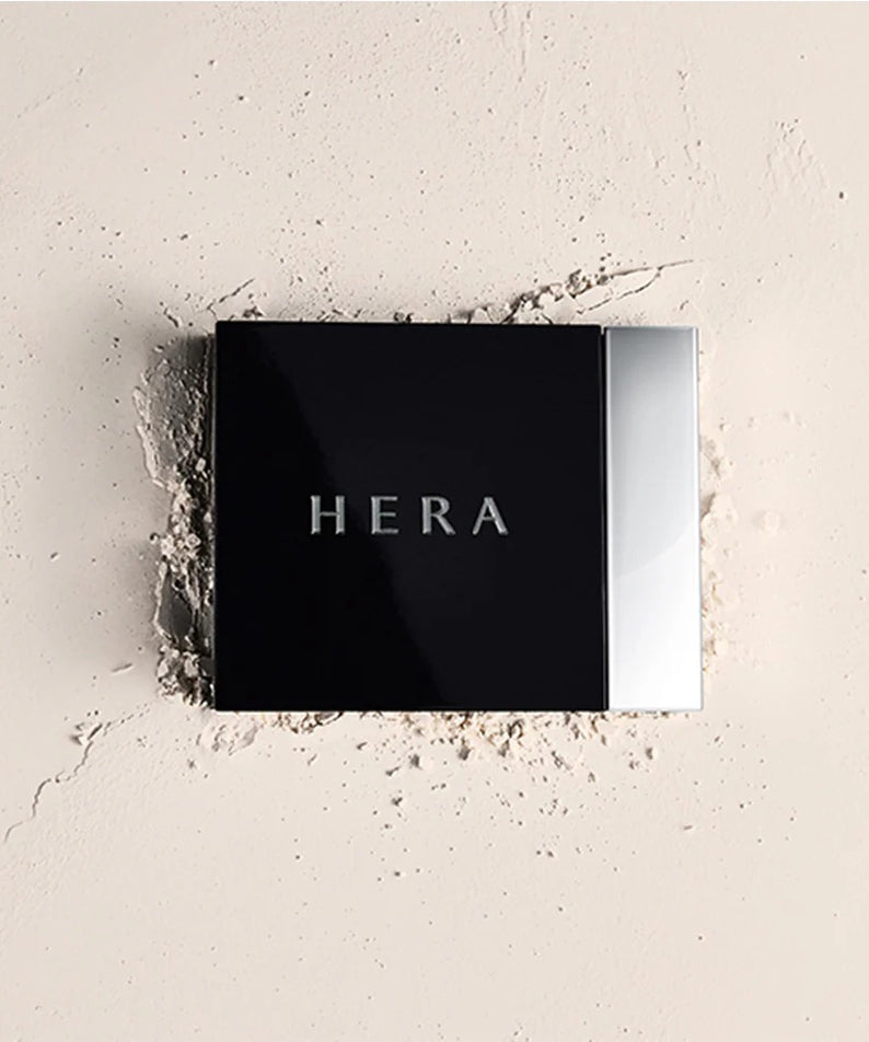 Boîtier du HERA Airy Powder Primer 8.5g sur fond de poudre diffusée.