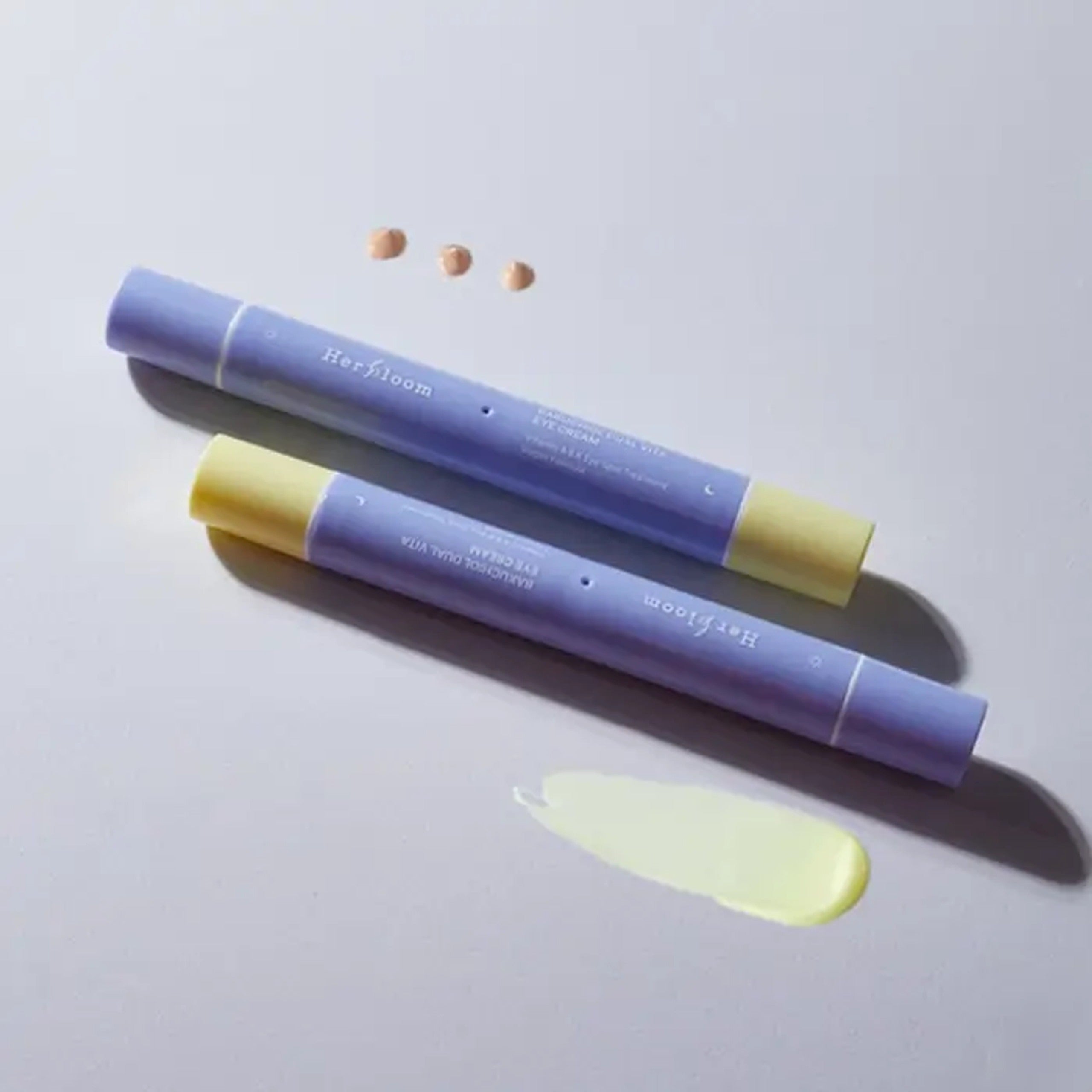 Décrit l’image en français : HERBLOOM  Bakuchiol Dual Vita Eye Cream, tube bleu-violet, texture crème près d’un fond clair.
