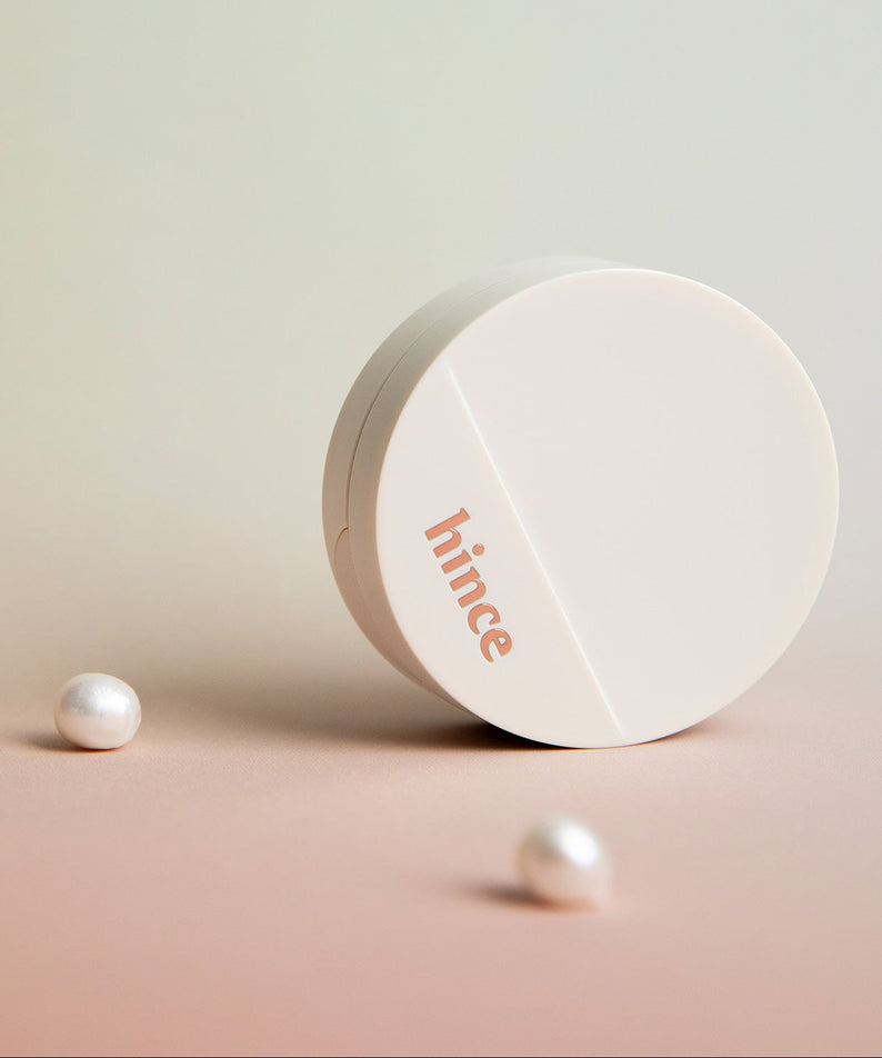 HINCE  Second Skin Glow Cushion de HINCE, boîtier blanc sur fond nude avec perles effet premium, maquillage lumineux