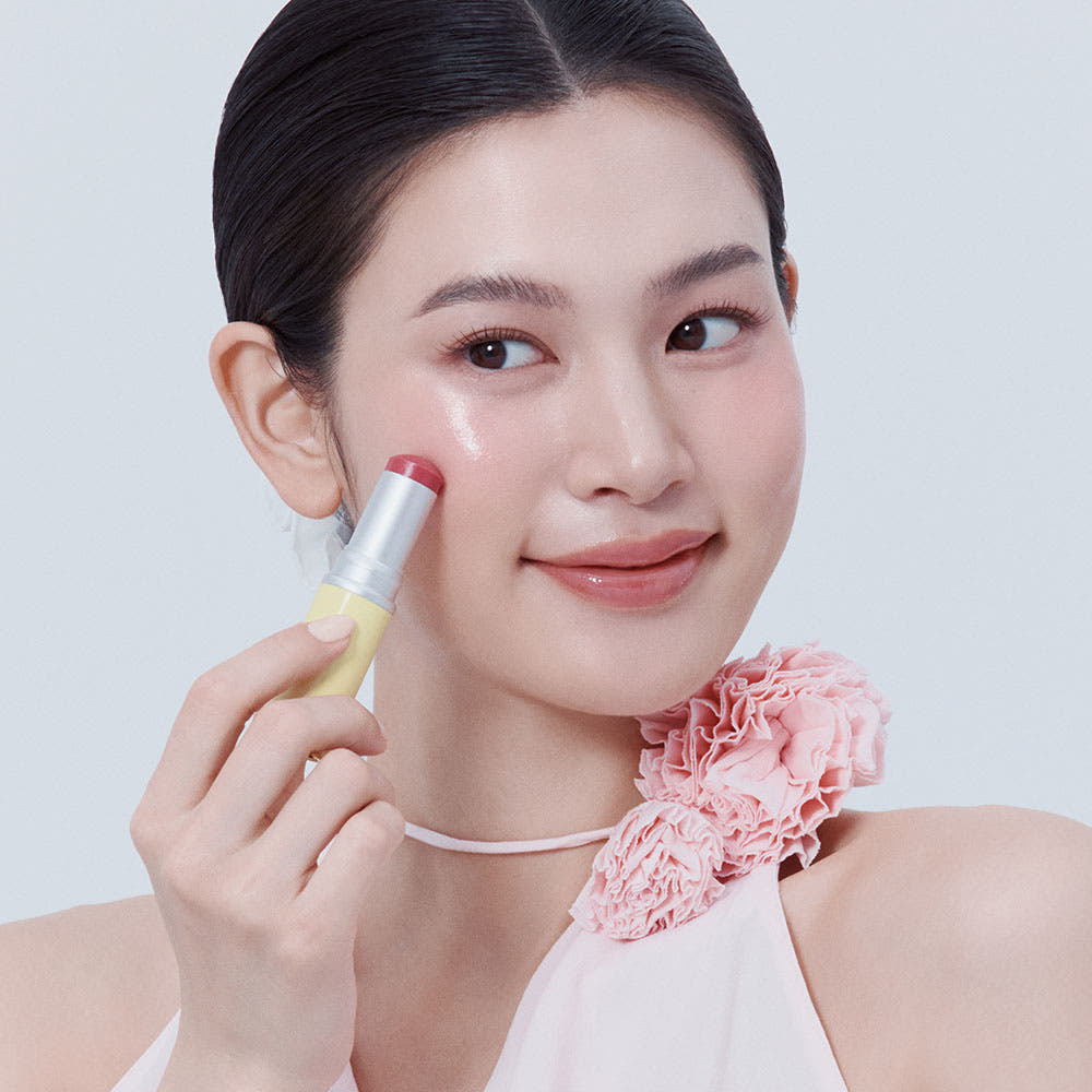 HINCE True Dimension Radiance Balm 10g : femme applique le baume sur fond clair, flacon HINCE visible, fleurs roses en décor.