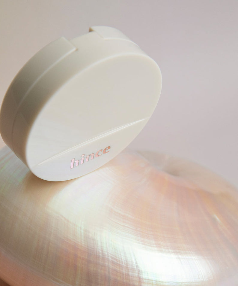 Coussin HINCE Second Skin Glow Cushion posé sur une coquille irisée, reflétant une lueur subtile et élégante.