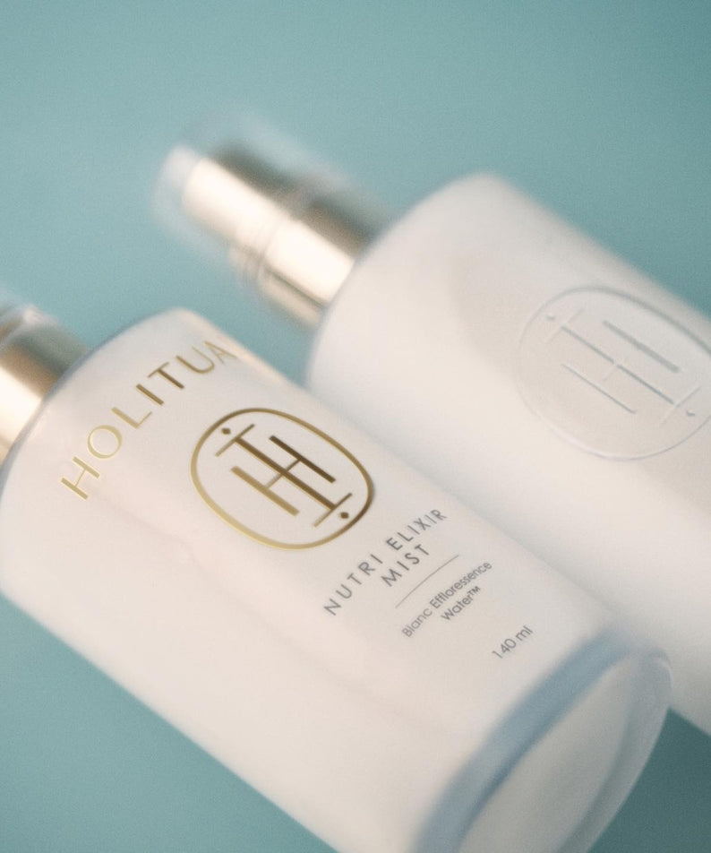 Flacons de HOLITUAL Elixir Mist 140ml posés sur une surface bleue, capturant l'essence du soin beauté.