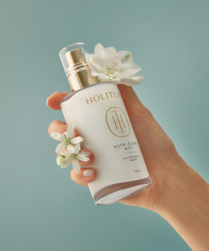 Flacon de HOLITUAL Elixir Mist 140ml tenu à la main avec des fleurs blanches, sur fond bleu, produit cosmétique élégant.