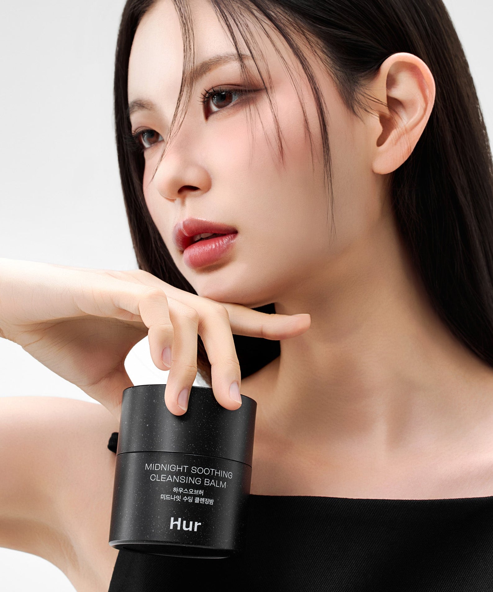 Décris l’image en français : HOUSE OF HUR Midnight Soothing Cleansing Balm HUR, femme en soin, pot noir sur fond clair.