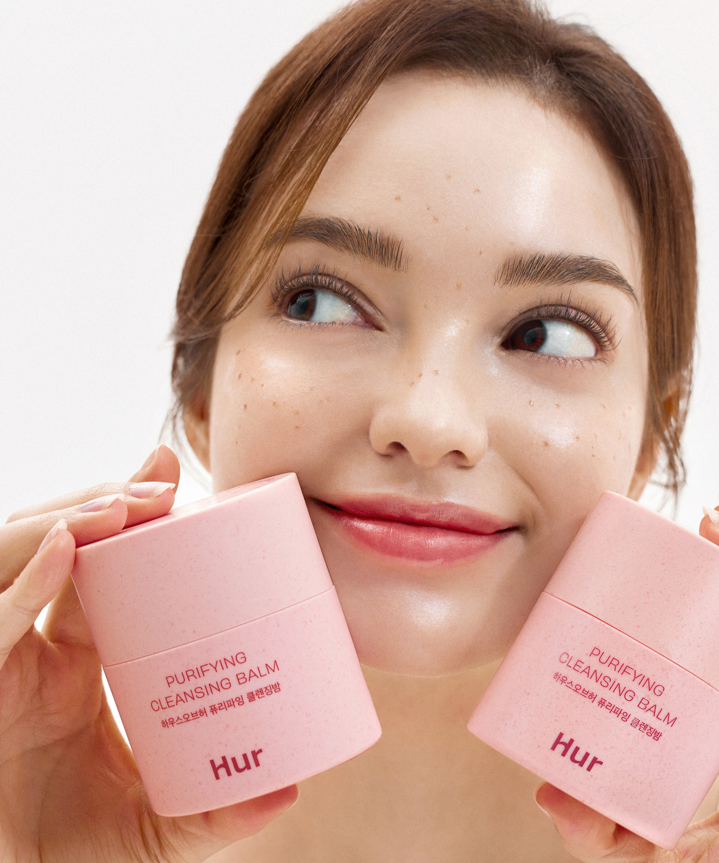 Décris l'image en français : HOUSE OF HUR Purifying Cleansing Balm, femme tenant deux baumes roses contre fond blanc.
