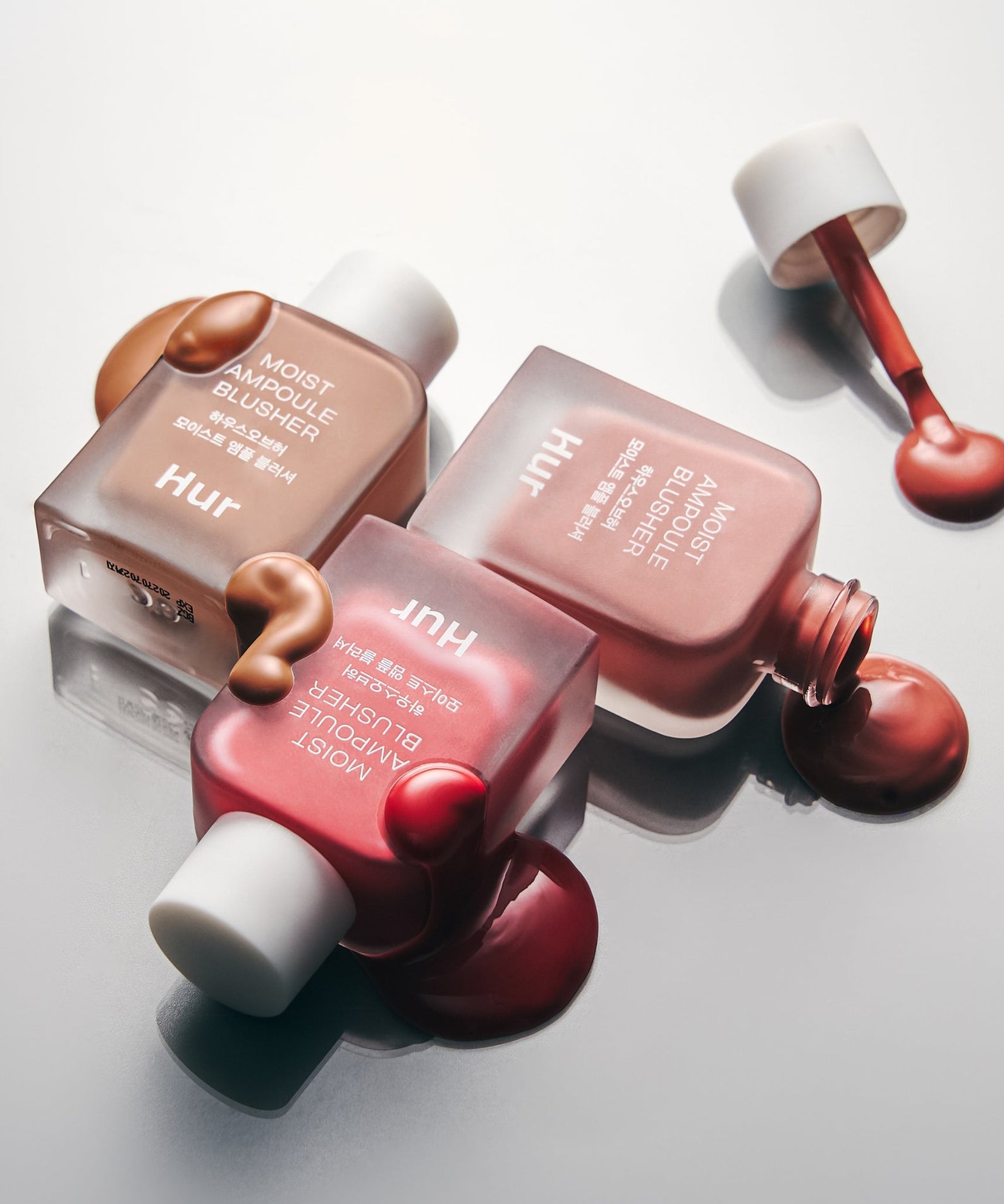 Photo de trois flacons de HOUSE OF HUR Moist Ampoule Blusher 10ml avec blush liquide renversé sur une surface blanche élégante.