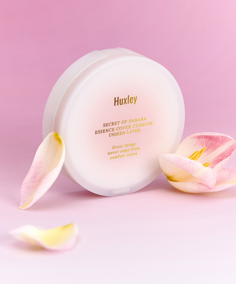 Coussin de teint HUXLEY Essence Cover Cushion Unseen Layer sur fond rose avec pétales, hydratant et couvrant pour une peau éclatante.