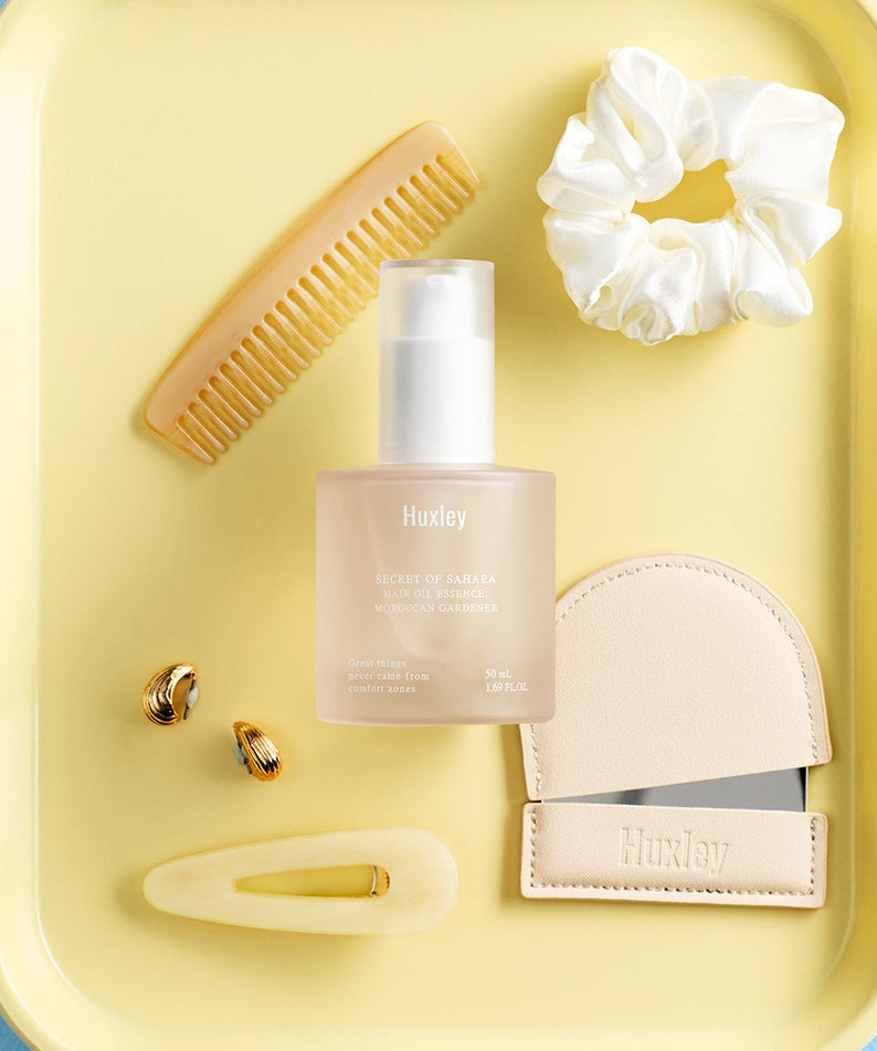 Décris l'image en français : flacon HUXLEY Hair Oil Essence Moroccan Gardener 50ml, marque HUXLEY, sur fond jaune avec accessoires.