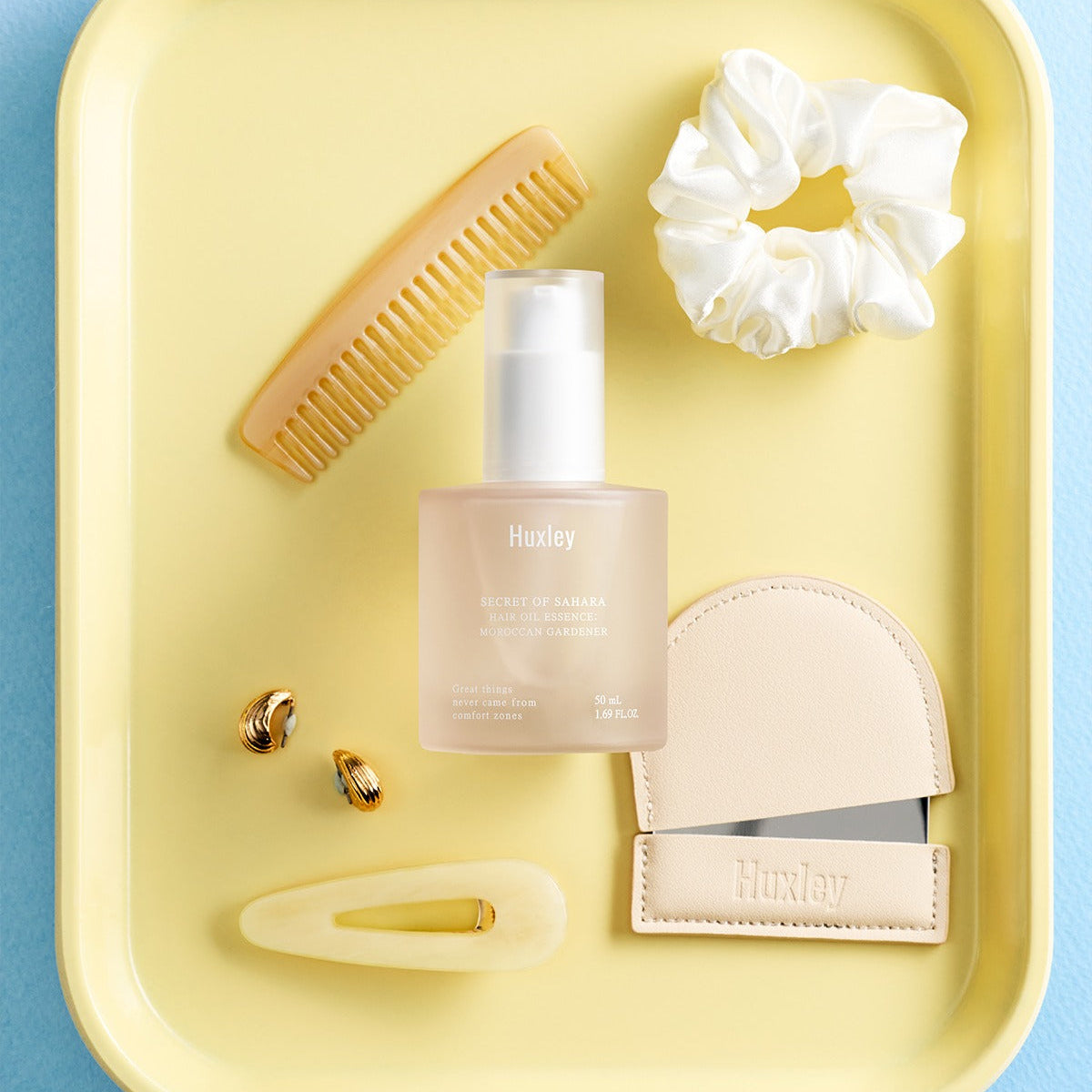 Décris l'image en français : flacon HUXLEY Hair Oil Essence Moroccan Gardener 50ml, marque HUXLEY, sur fond jaune avec accessoires.