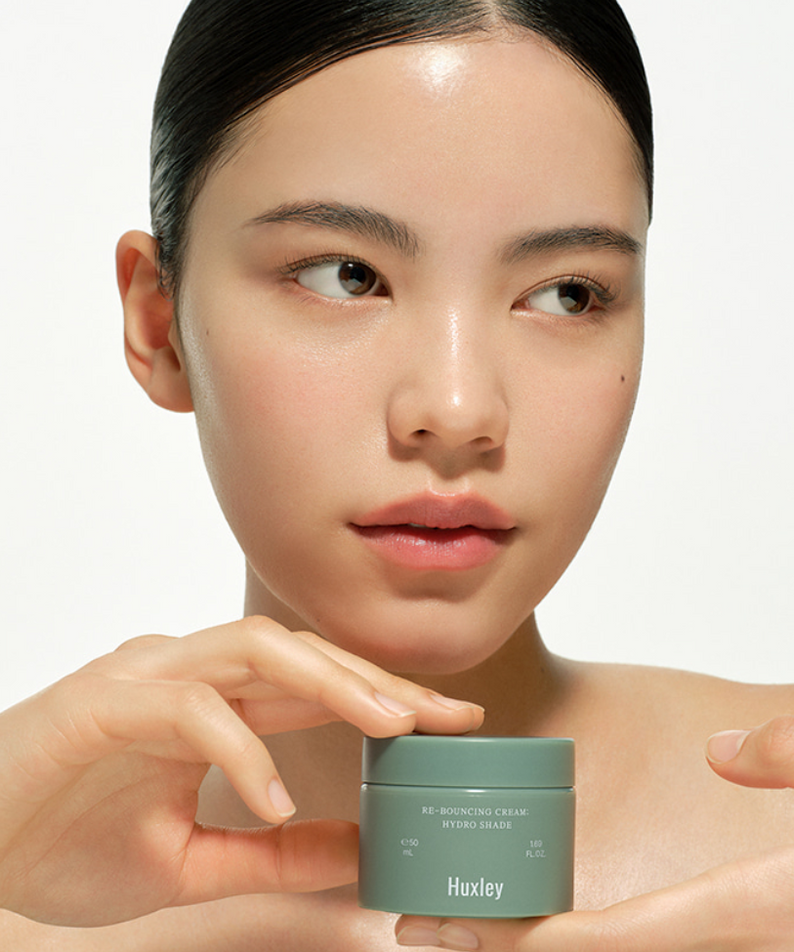 HUXLEY Rebouncing Cream Hydro Shade 50ml : crème hydratante sur une peau, pot vert près du visage (portrait beauté, marque HUXLEY).