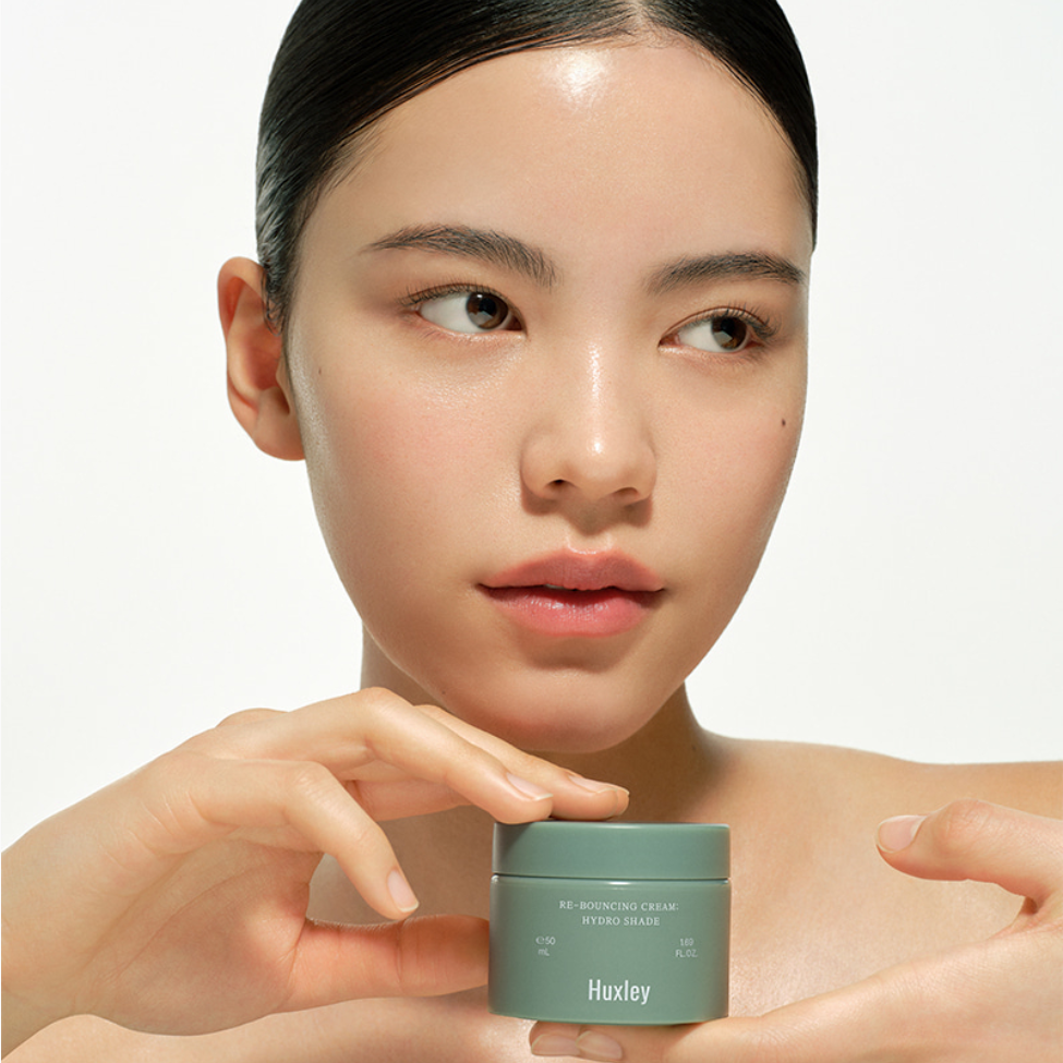 HUXLEY Rebouncing Cream Hydro Shade 50ml : crème hydratante sur une peau, pot vert près du visage (portrait beauté, marque HUXLEY).