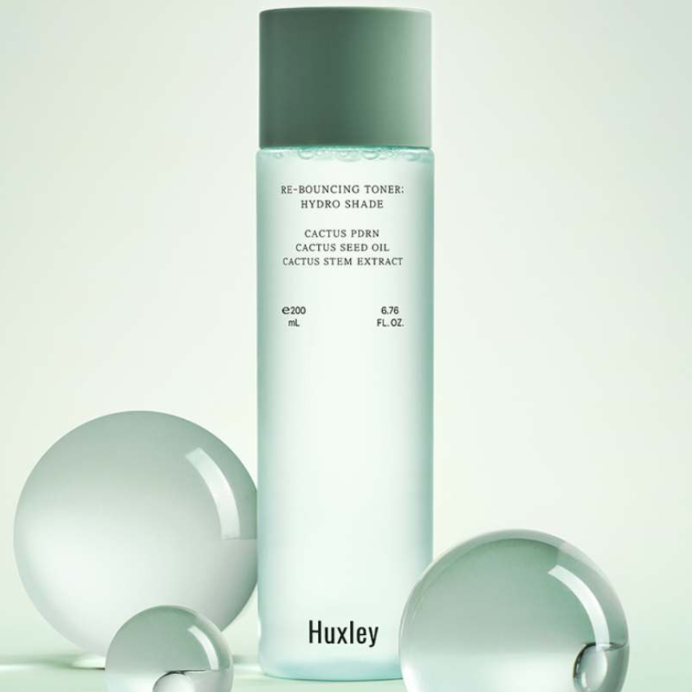 HUXLEY Rebouncing Toner Hydro Shade 200ml, flacon vert sur fond clair avec produits beauté, décor minimaliste.