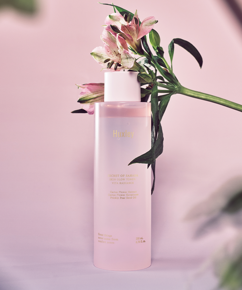 HUXLEY Skin Glow Toner Vita Radiance, toner hydratant et éclaircissant avec flacon rose et fleurs en arrière-plan.