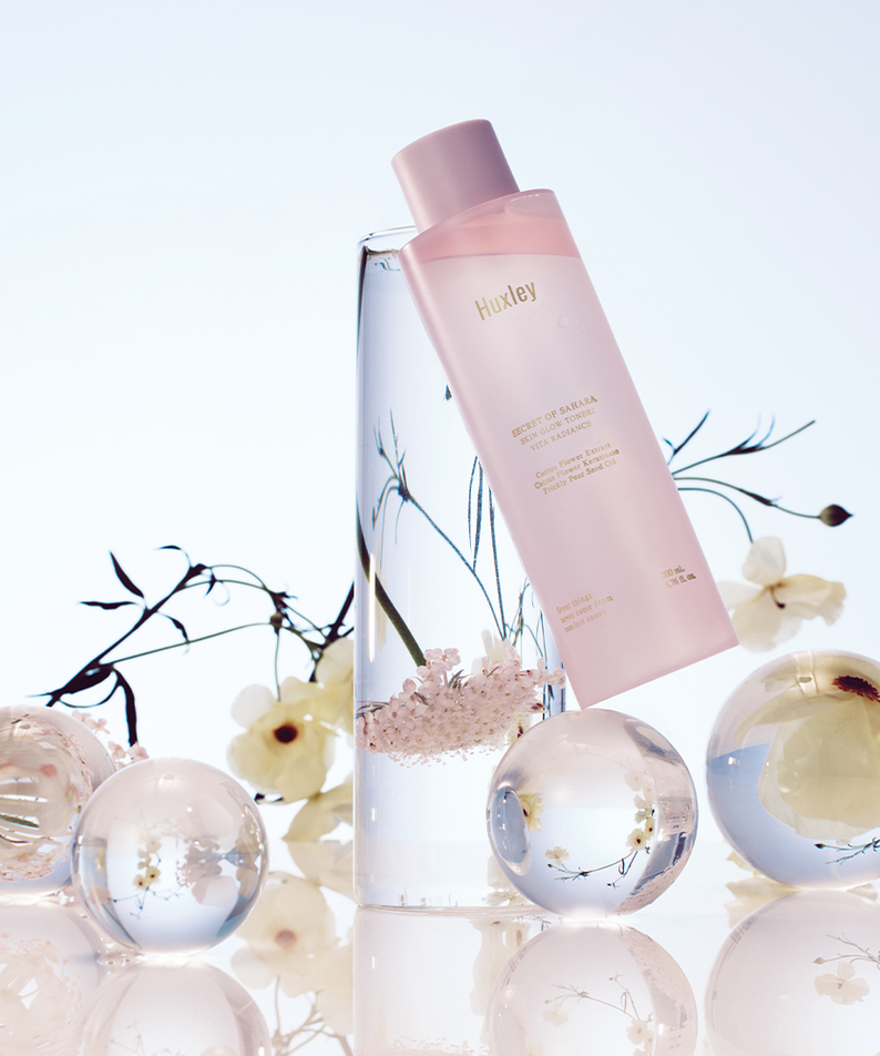 Flacon de HUXLEY Skin Glow Toner Vita Radiance entouré de fleurs et de sphères en verre, ambiance luxueuse et rafraîchissante.