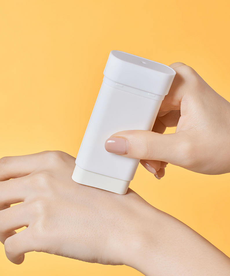 Main tenant ILLIYOON Daily Weaponless Sun Stick 20g, marque sur tube blanc, fond jaune ensoleillé.
