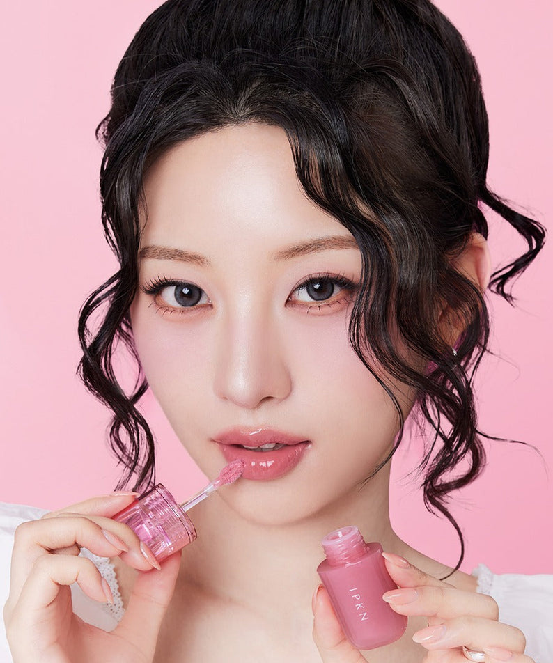 Femme appliquant IPKN Bubble Jam Gloss, un brillant à lèvres rose, avec fond rose vif. Parfait pour une finition brillante et élégante.