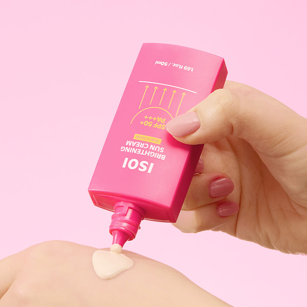 Main tenant ISOI Brightening Sun Cream SPF 50+ PA +++ 50ml sur peau, crème solaire éclaircissante, fond rose pastel.