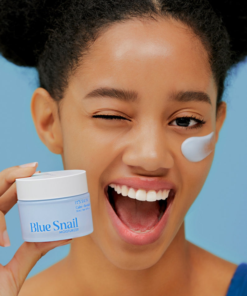 Jeune femme souriante tenant IT'S SKIN Blue Snail Cream 50ml avec une touche de crème sur la joue, fond bleu.