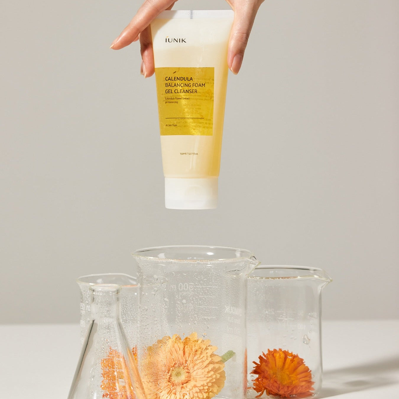 IUNIK Calendula Balancing Foam Gel Cleanser 150ml, flacon tenu en main, fleurs de calendula sur fond clair