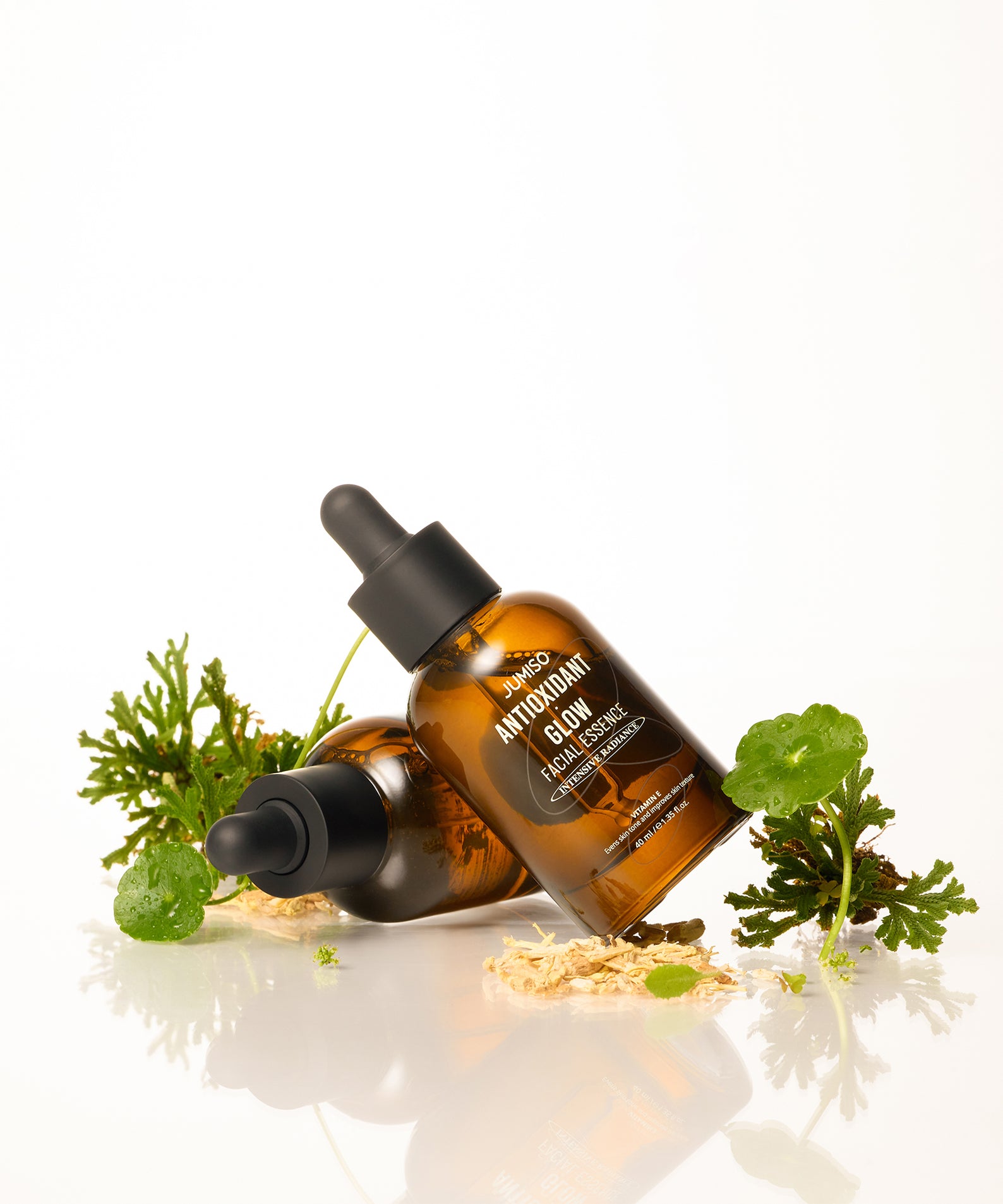 Flacons de JUMISO Antioxidant Glow Facial Essence posés sur un fond blanc avec des plantes vertes pour un éclat naturel.