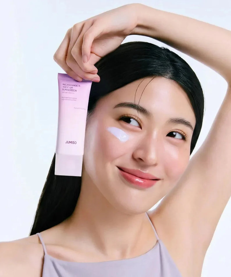 Décris l'image en français : JUMISO  Niacinamide 3 Plus Tone-Up Sunscreen 40ml, femme l'applique, flacon rose JUMISO sur fond clair.