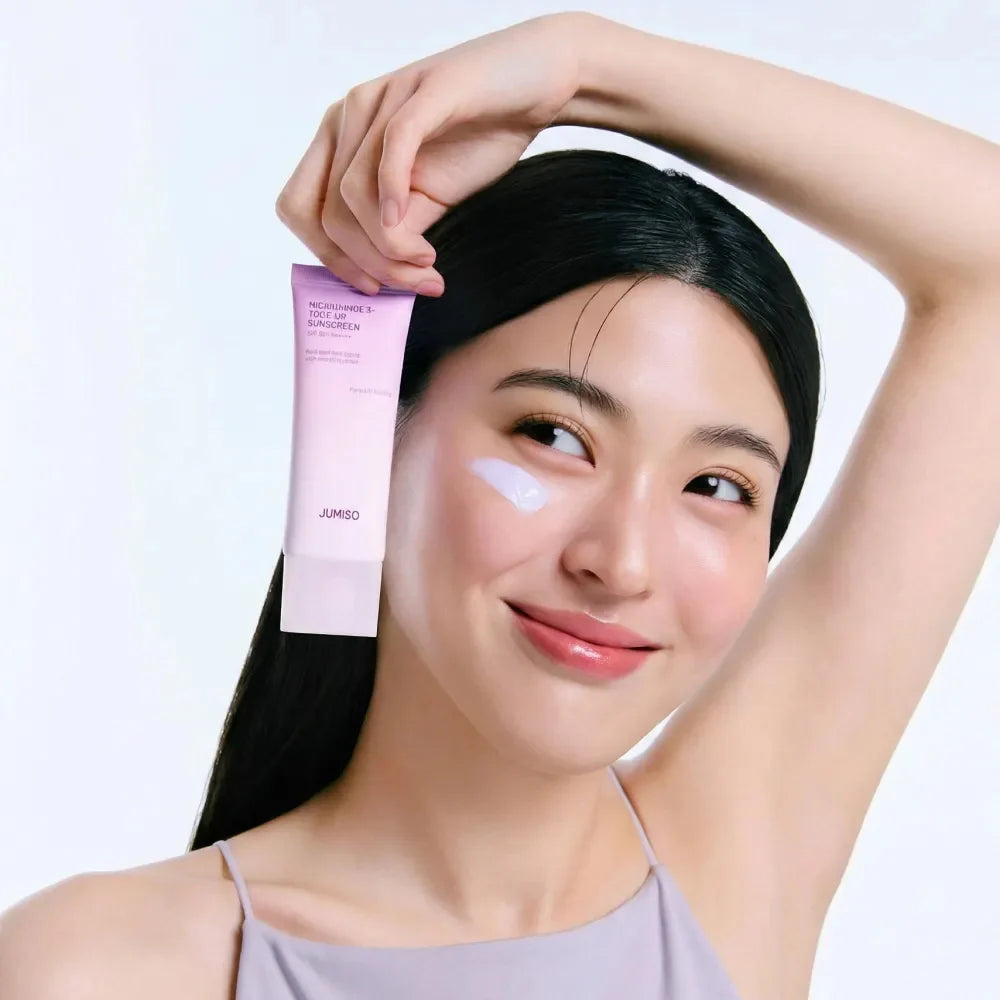 Décris l'image en français : JUMISO  Niacinamide 3 Plus Tone-Up Sunscreen 40ml, femme l'applique, flacon rose JUMISO sur fond clair.