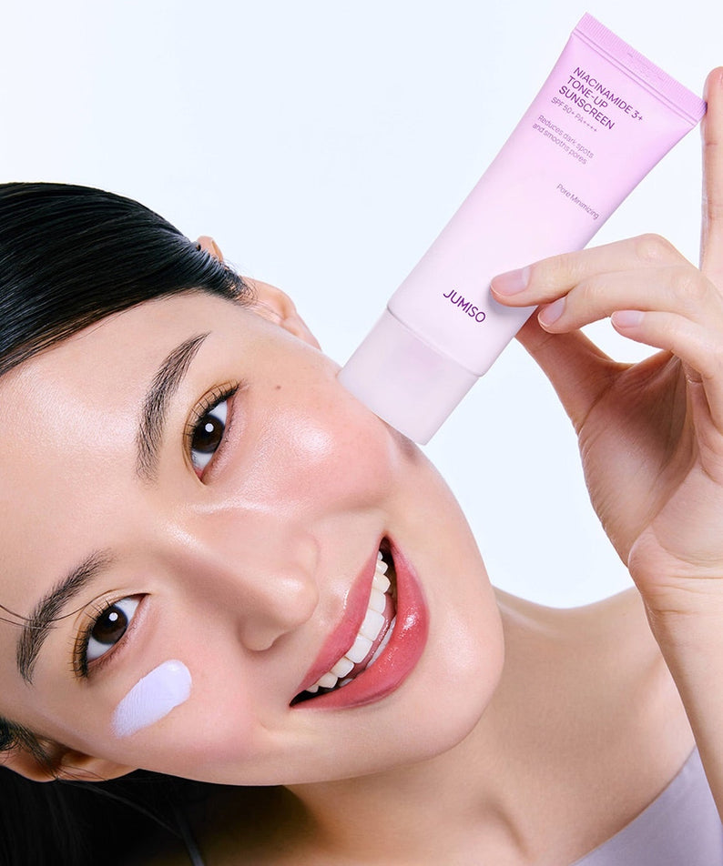 Décris l'image en français : JUMISO  Niacinamide 3 Plus Tone-Up Sunscreen 40ml, tube rose sur peau, main et visage.