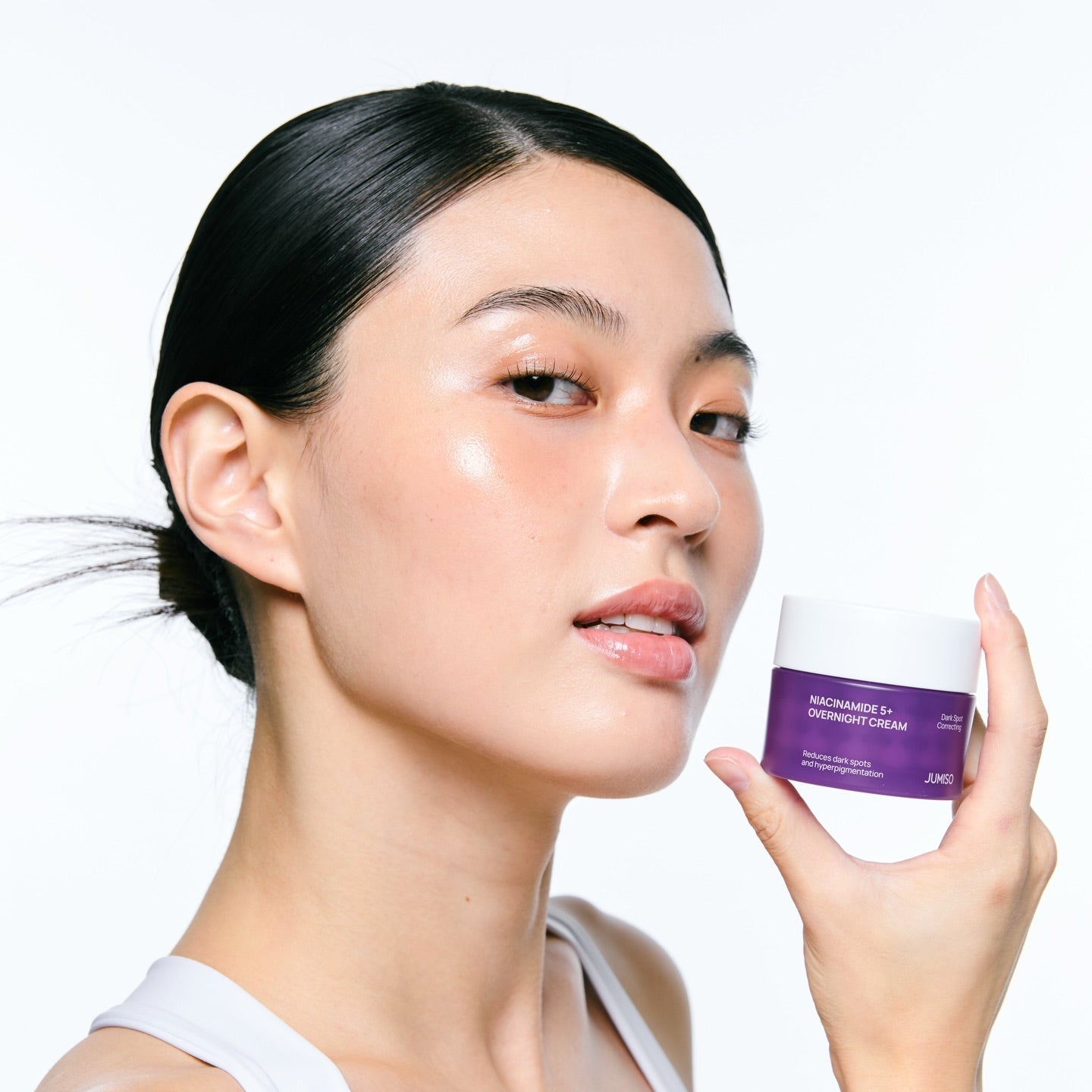 Décris l'image en français : JUMISO  Niacinamide 5 Plus Overnight Cream 80ml, femme tenant le pot, studio skincare.