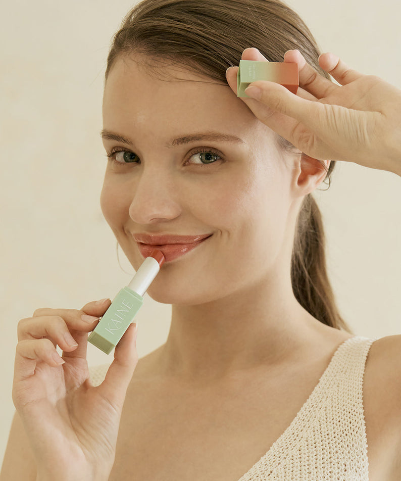 Femme souriante appliquant le KAINE Glow Melting Lip Balm, produit hydratant pour lèvres éclatantes et douces.