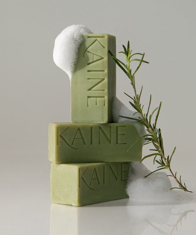 Trois savons empilés avec mousse et une branche de romarin, KAINE Rosemary Acne Mildly Acidic Cleansing Bar 100g illustré.
