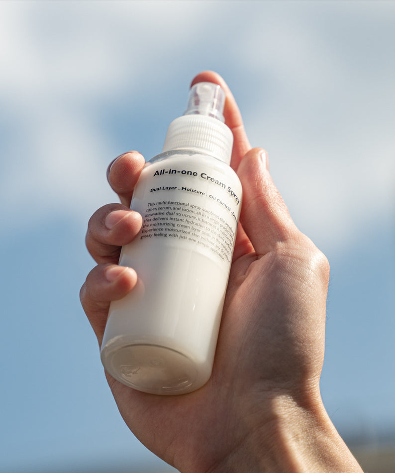 LAZY SOCIETY All-in-One Cream Spray 120ml : flacon crème all-in-one tenu à la main, fond ciel bleu, vue rapprochée.