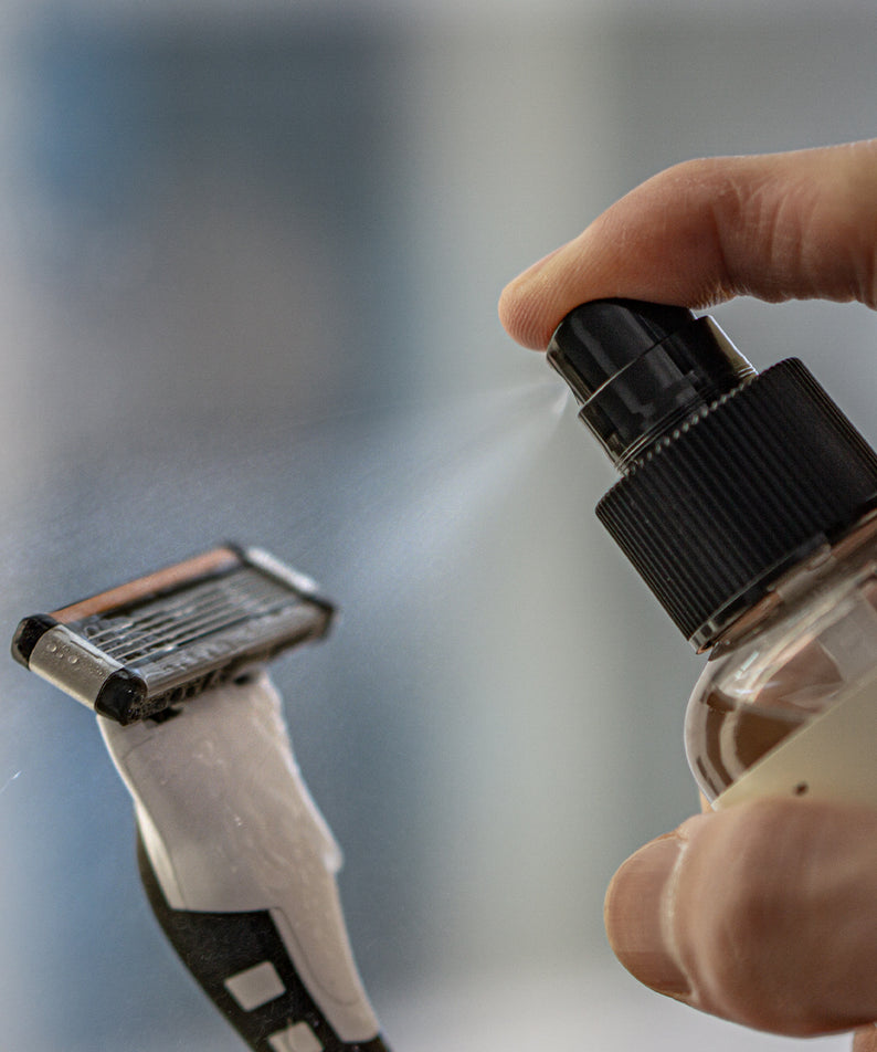 LAZY SOCIETY Razor Blade Sanitizer 120ml : main tenant le spray, rasoirs en arrière-plan, contexte de désinfection.