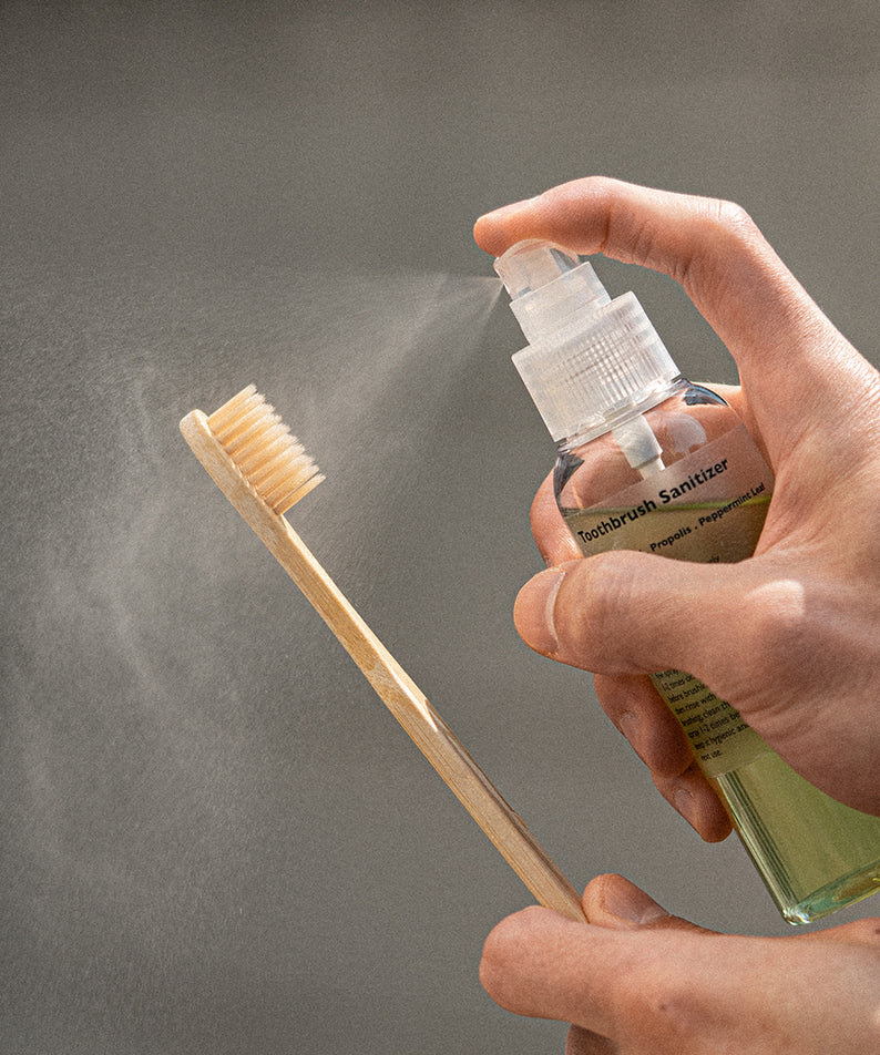 Décris l'image en français : LAZY SOCIETY Toothbrush Sanitizer 100ml, main vaporisant sur brosse, fond gris, marque visible.