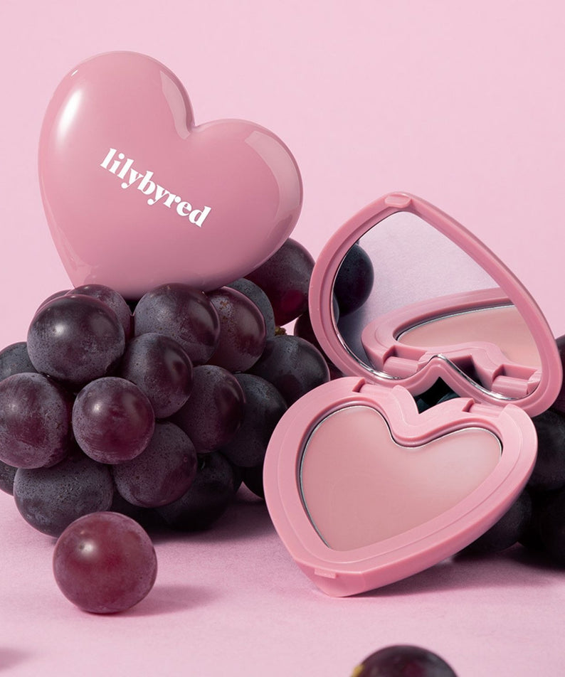 LILYBYRED Love Beam Cheek Balm en forme de cœur avec miroir sur fond rose, entouré de raisins noirs.