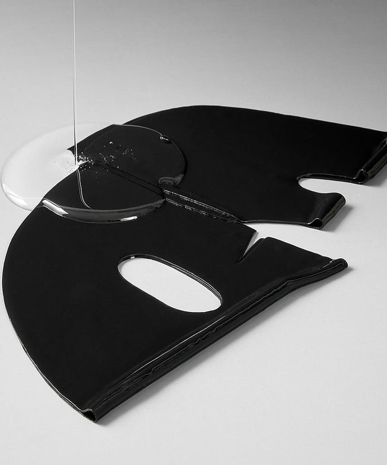 Feuille de masque noire du MARY&MAY Vegan Collagen Charcoal PDRN Gel Mask (4 Sheets) avec solution liquide versée dessus, vue artistique.