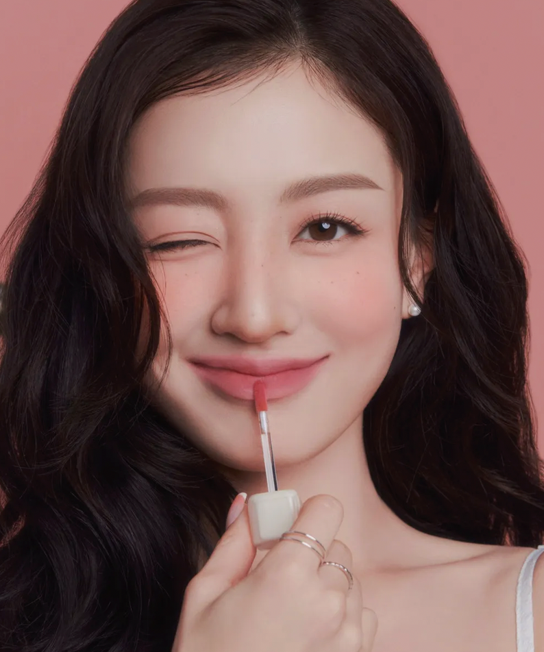 Femme appliquant MILK TOUCH Spread Fit Blur Tint, maquillage naturel et élégant, fond rose, rouge à lèvres hydratant en France.