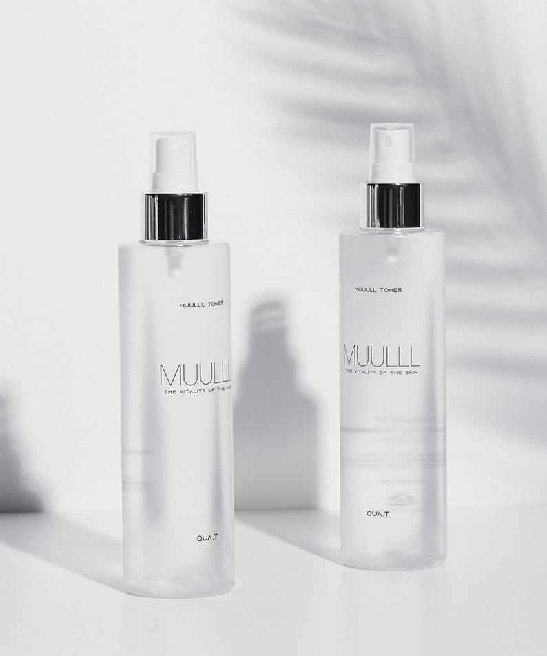 Deux bouteilles de QUA-T Toner 200ml posées sur une surface blanche élégante, produisant une ambiance sophistiquée. Packaging minimaliste chic.