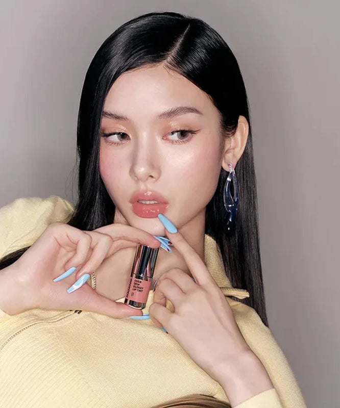 NAMING Overdew Glossy Lip Tint, femme aux lèvres brillantes tenant le produit sur fond neutre, maquillage glam