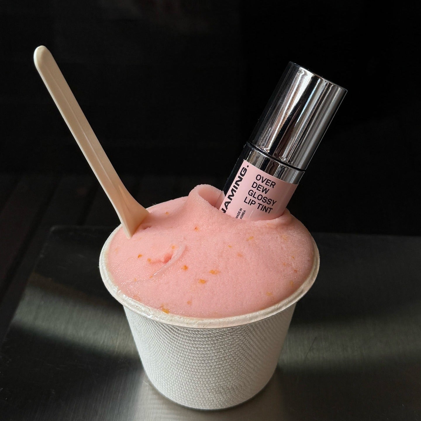 NAMING Overdew Glossy Lip Tint de la marque NAMING, sur fond noir, présenté sur une glace rose en studio