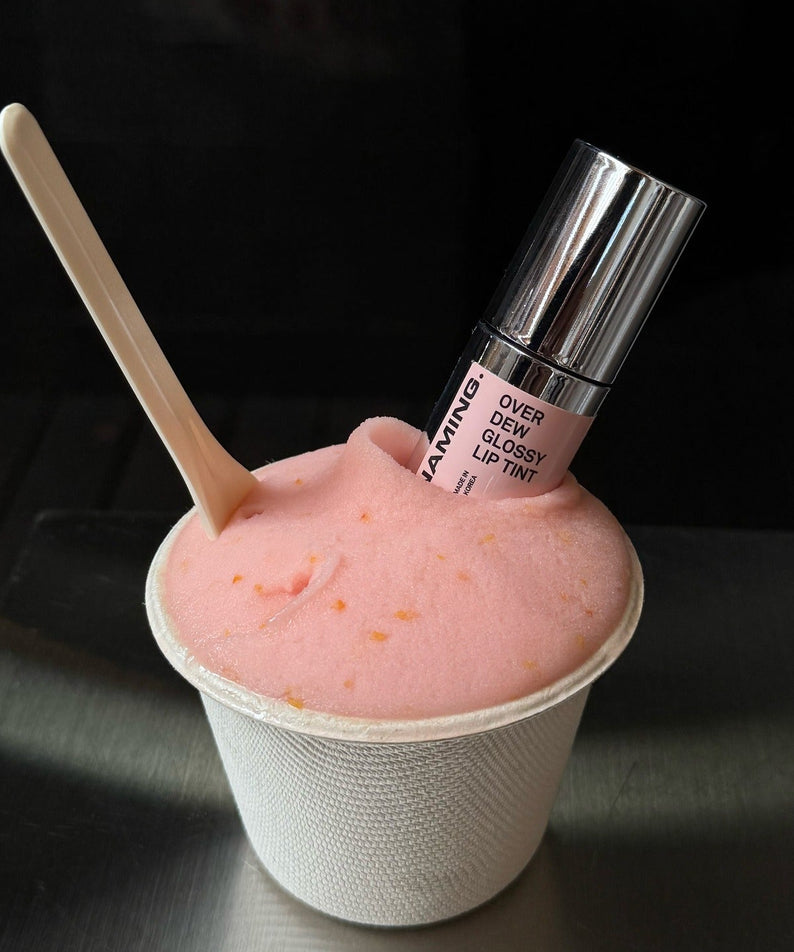 NAMING Overdew Glossy Lip Tint posé dans une glace rose, présentant le rouge à lèvres brillant pour une application élégante.