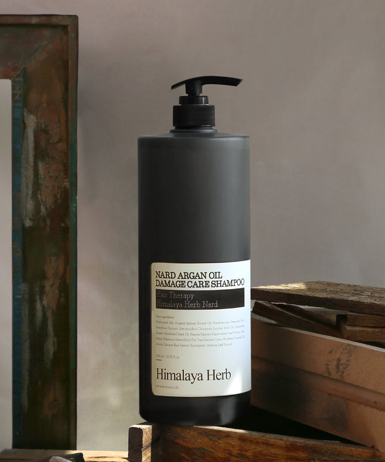 Décris l'image en français : NARD Argan Oil Damage Care Shampoo 1500ml de NARD, flacon noir sur bois, lumière naturelle.