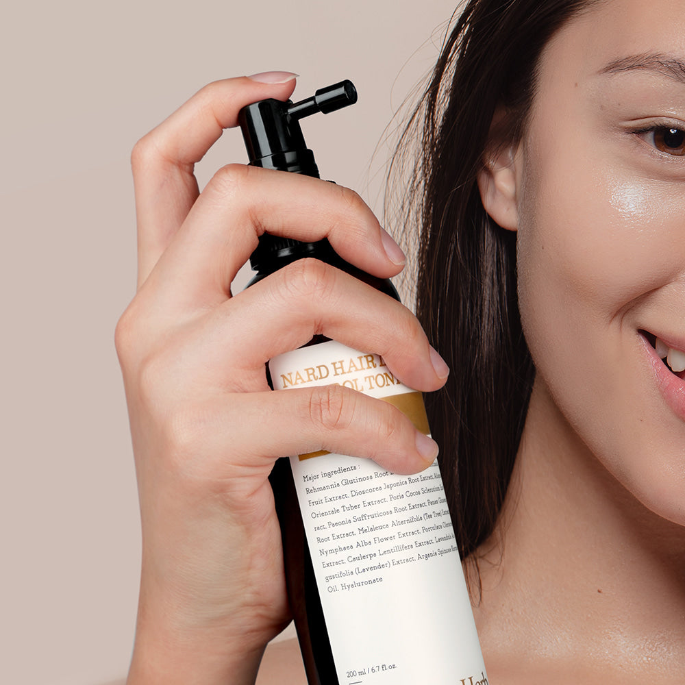 Décris l'image en français : NARD Hair Loss Relief Hair Tonic 200ml de la marque NARD, appliqué sur le visage, main tenant le flacon.