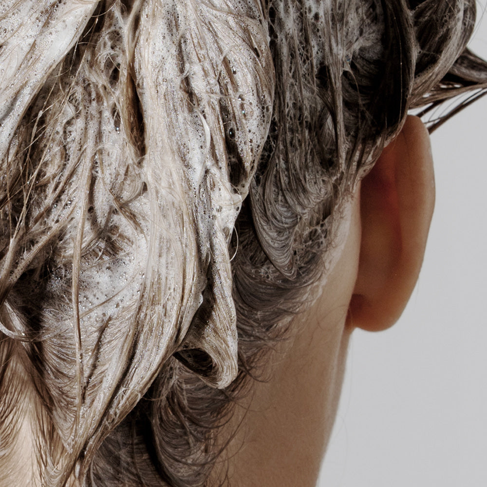 Décris l'image en français : NARD Repairing Shampoo 1500ml de la marque NARD, cheveux avec mousse sous douche blanche.