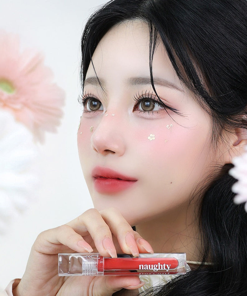 NAUGHTY PERCENT Matte Bloom Tint de la marque NAUGHTY PERCENT, maquillage mat sur modèle devant fleurs, flacon rouge en gros plan.