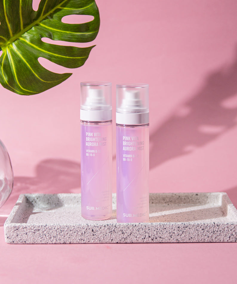 Deux bouteilles de NEOGEN Pink Vita Aurora Mist 115ml posées sur un plateau texturé avec feuille verte en fond, ambiance rose.