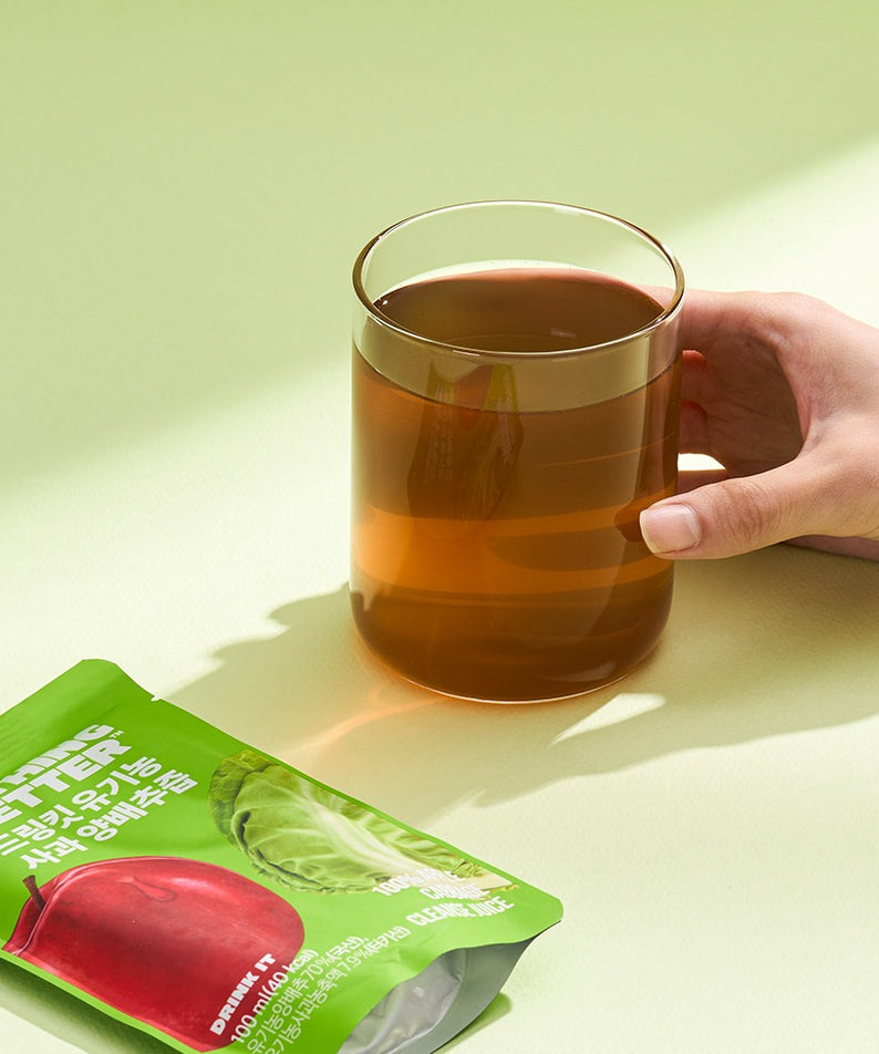 Décris l'image en français : NOTHING BETTER Drinkit Organic Applr Cabbage Juice 100ml 24 Packets, mains tient mug, sachet vert.