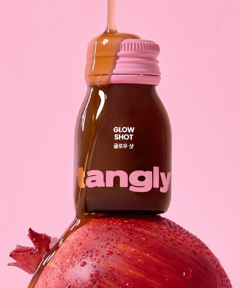 NOTHING BETTER Tangly Glow Shot 7 Bottles posé sur une grenade, avec un fond rose vibrant.
